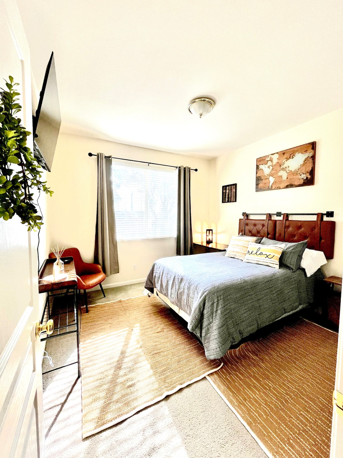 A well-lit bedroom is furnished with a spacious bed featuring a dark wood headboard. A desk and chair are positioned next to the bed, and the window is dressed with light-filtering curtains. A small rug lies on the carpeted floor, enhancing the cozy atmosphere.