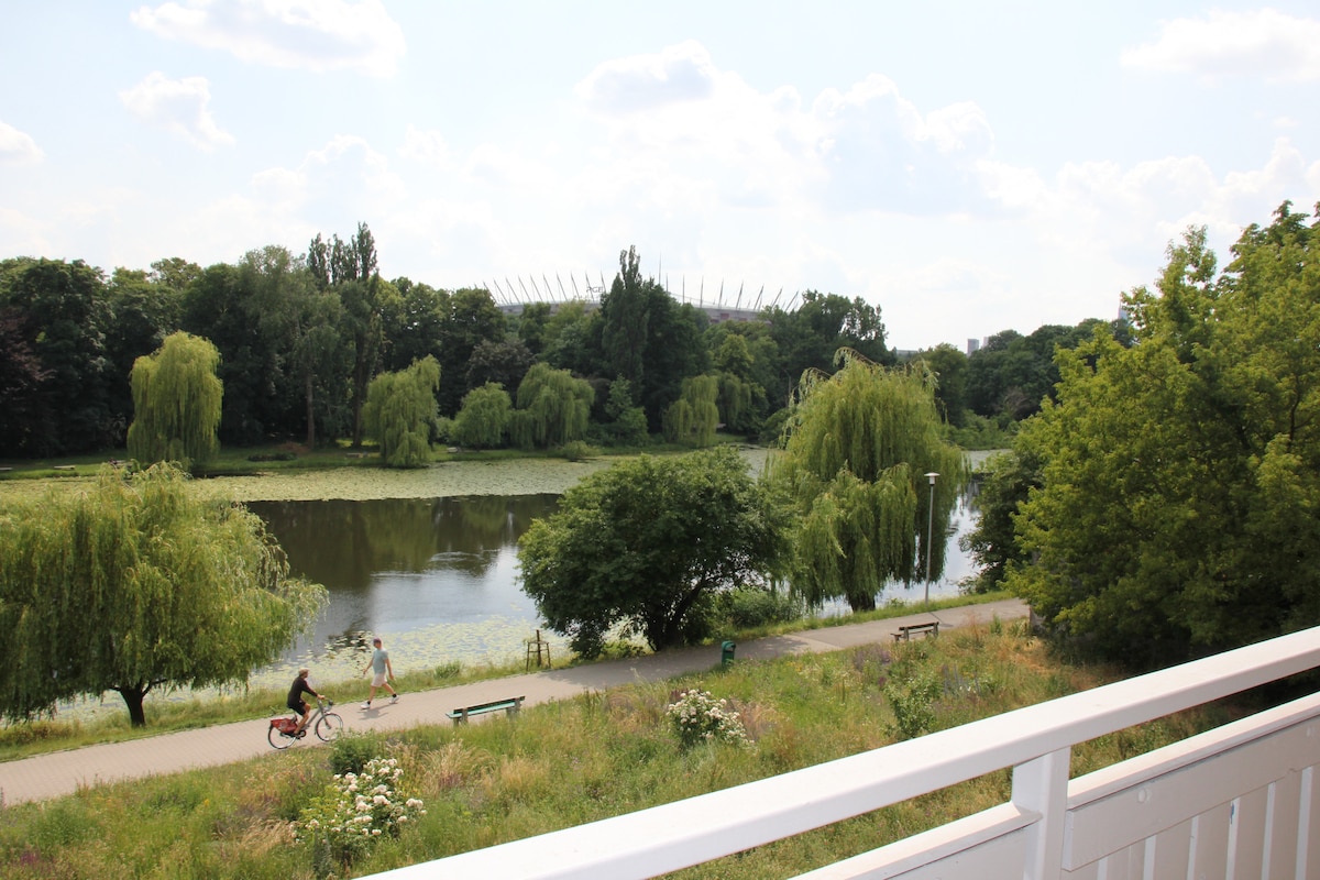 A serene view overlooks a lush green park with a tranquil lake reflecting the trees. Bicyclists can be seen along a pathway, while benches are placed in the foreground for relaxation. The stadium is visible in the distance, framed by the natural landscape.