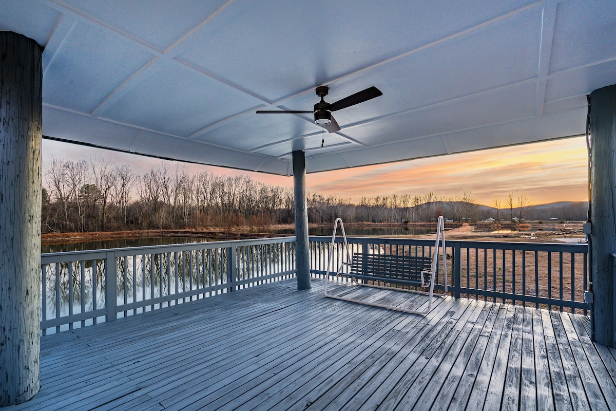 A spacious covered deck extends over the tranquil water, featuring a wooden swing and a ceiling fan. The expansive area provides scenic views of the surrounding landscape and reflects the soft colors of sunrise across the water's surface.