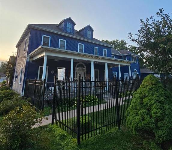 The historic exterior of The Wen Inn is showcased, featuring a striking blue facade and a spacious covered porch with white columns. A black wrought-iron fence outlines the property, with lush green landscaping and well-maintained garden areas visible in the foreground.