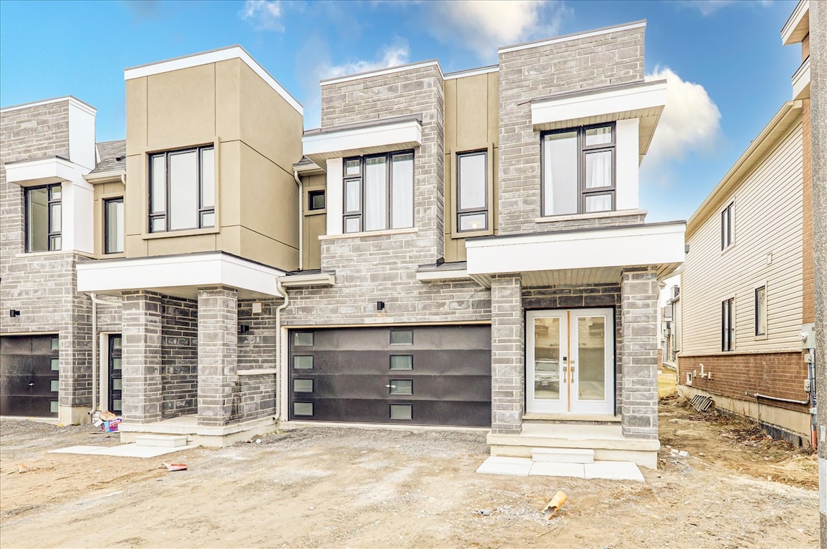 A modern two-story home features a unique façade of stone and smooth surface materials. The garage is equipped with a large, dark panel door. A clearly defined entrance is located at the front, leading to a well-designed entryway.