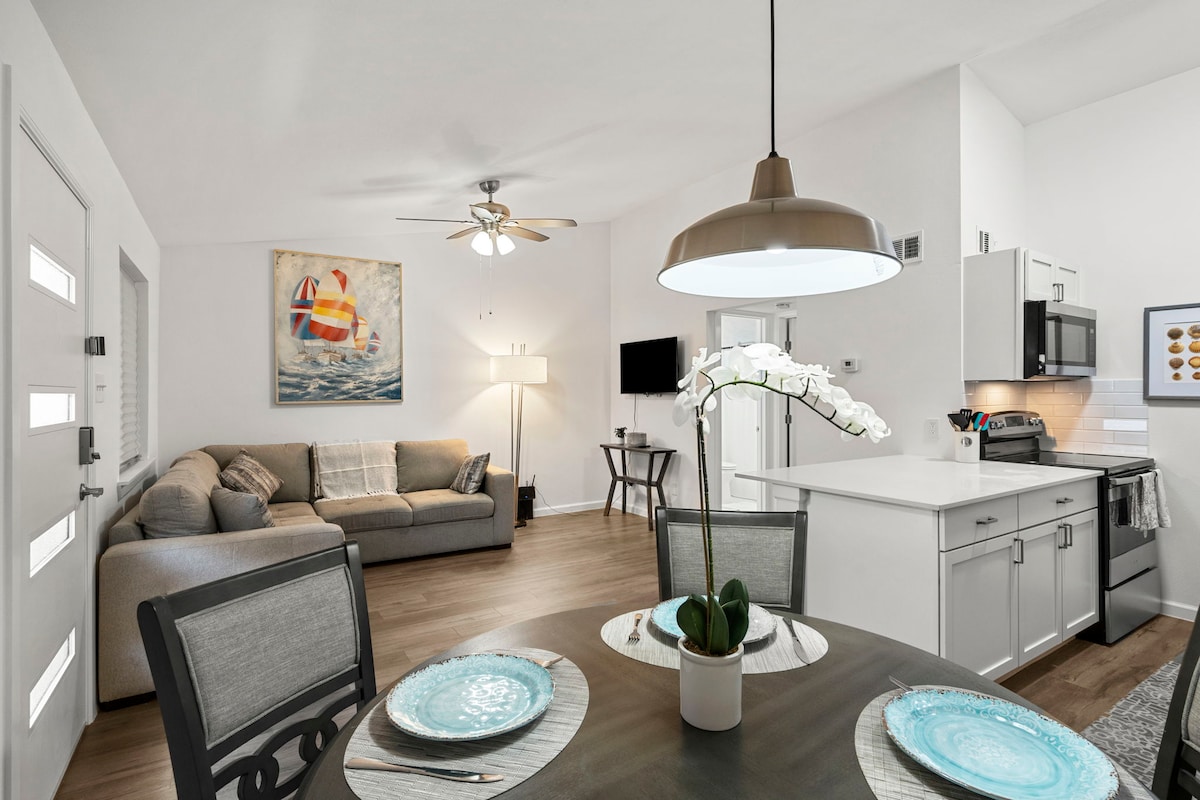 The bright living area features a modern couch, accompanied by a ceiling fan and light fixture. A dining table is set for four, displaying plates and a potted orchid. The kitchen showcases sleek, new appliances and a wall-mounted TV is visible from the seating area.