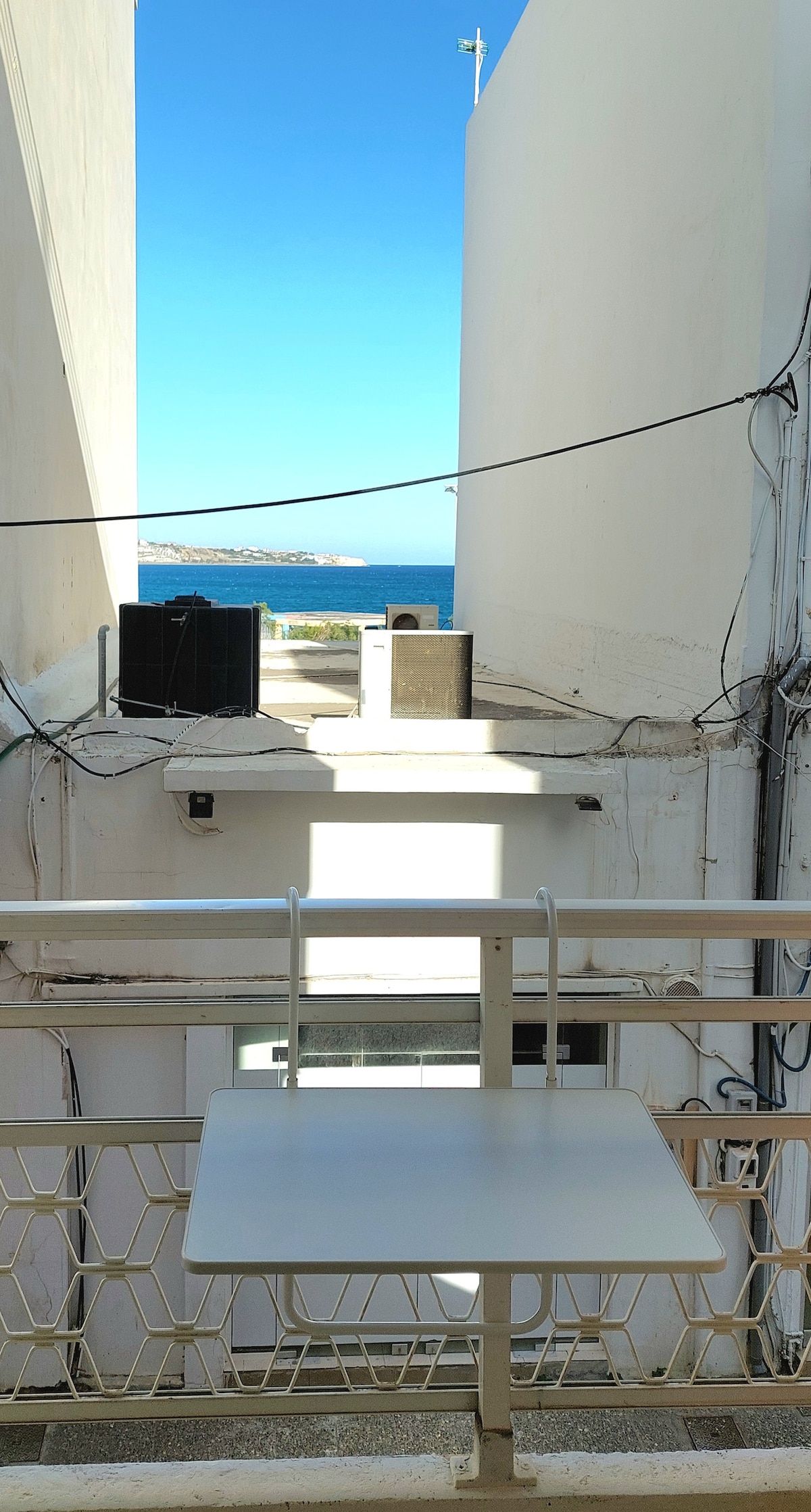 A balcony view is presented, showcasing light-colored walls and a small white table. Beyond the railing, a glimpse of the ocean is visible, framed by adjacent building walls. Clear blue skies are seen above, revealing a peaceful coastal atmosphere.