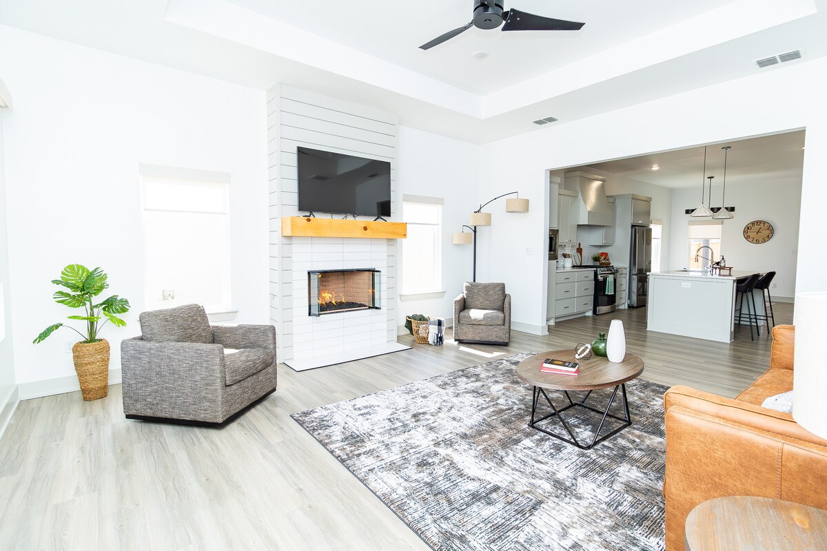 A spacious living area features two comfortable armchairs alongside a modern coffee table. A fireplace is set beneath a wall-mounted television, and a textured rug adds warmth to the space. The open concept connects seamlessly to the adjoining kitchen area.