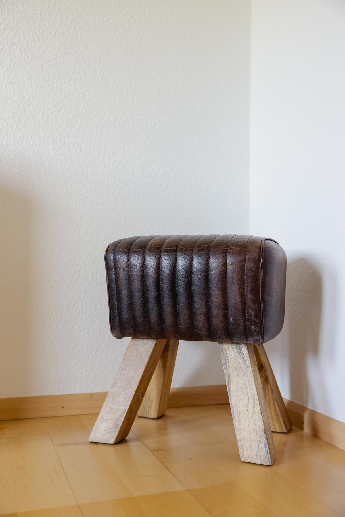 A round stool with a dark leather seat and vertical ribbed detailing is shown, supported by four wooden legs. The stool is placed on a hardwood floor against a light-colored wall, creating a simple yet functional addition to the space.