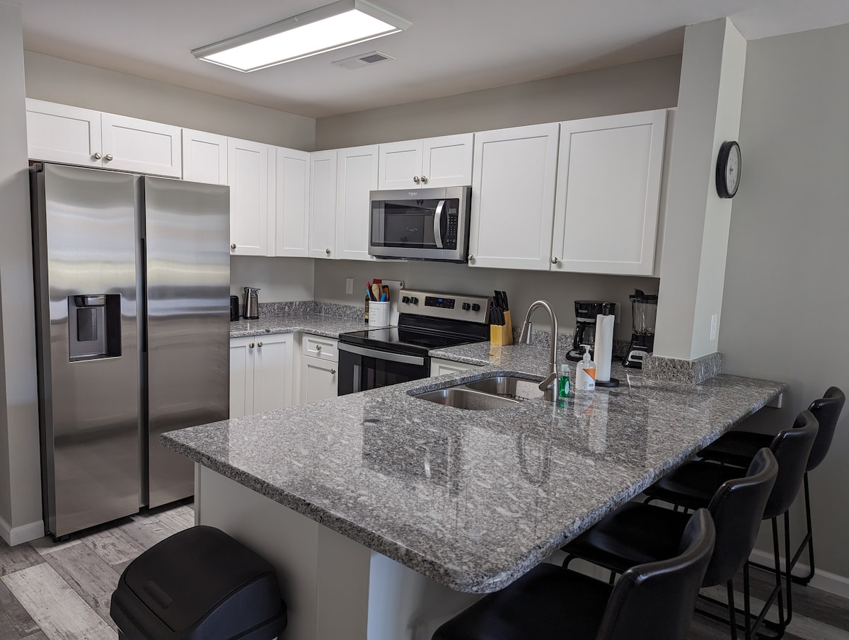 The kitchen features modern stainless steel appliances, including a refrigerator and microwave, set against sleek white cabinetry. A spacious island with seating for four creates a functional layout, complemented by a subtle gray countertop and ample natural light from the overhead fixture.