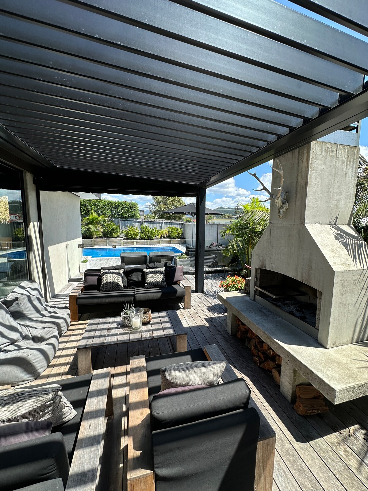 An outdoor seating area is situated under a sleek pergola, featuring comfortable black sofas adorned with decorative cushions. A wooden coffee table holds a small potted plant. In the background, a pool reflects the clear blue sky, while greenery and an outdoor fireplace enhance the space.