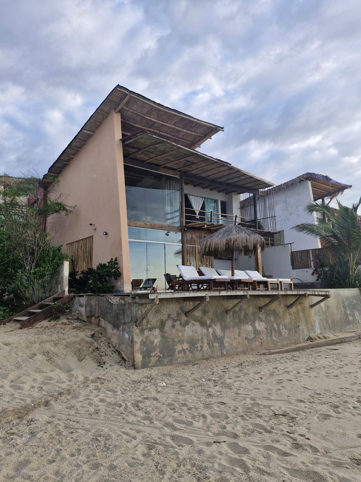 A beachfront house is showcased with a sandy area in the foreground. The structure has large glass walls, allowing for natural light. A covered terrace with seating is visible on the upper floor, complemented by a thatched roof. Surrounding greenery adds a touch of nature.