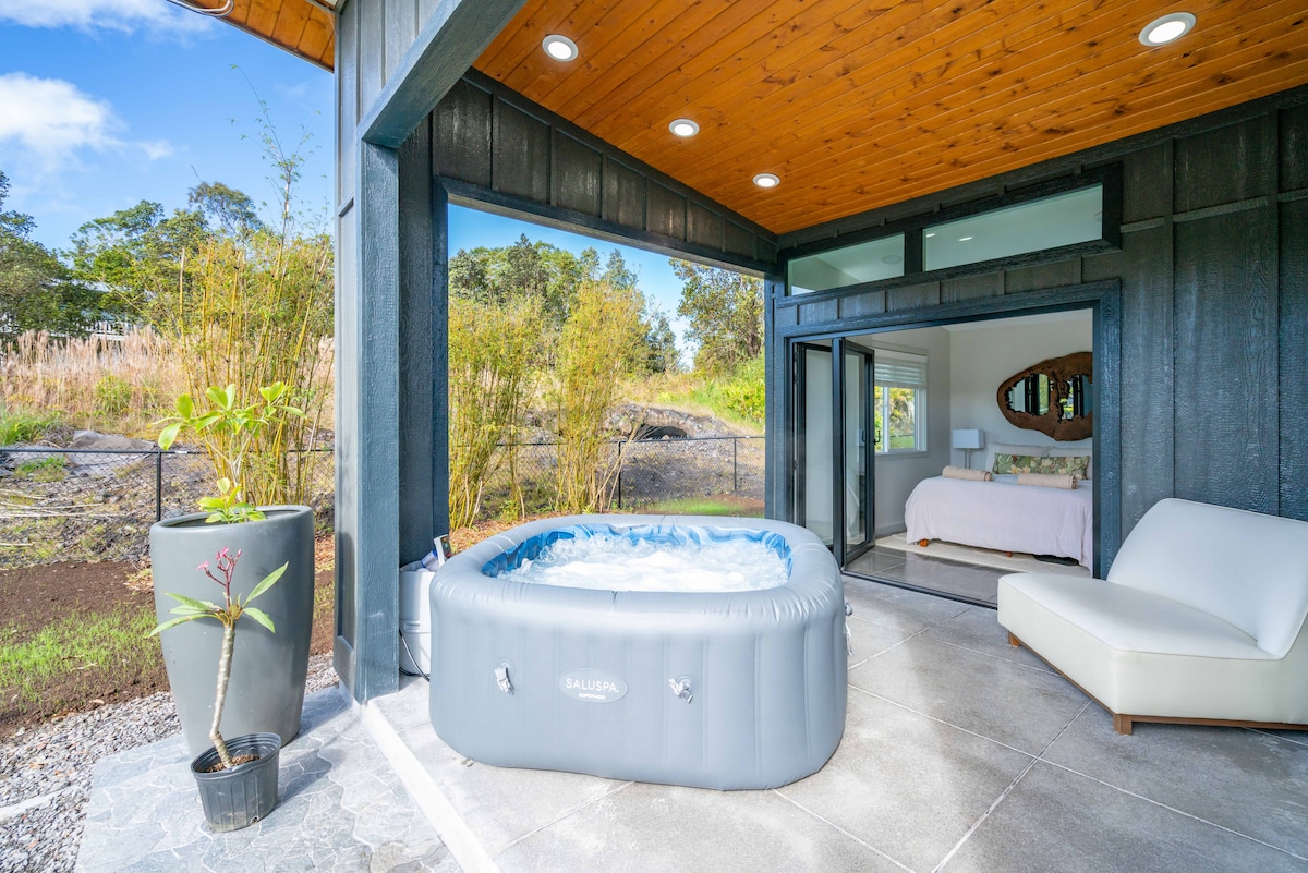 An outdoor jacuzzi is positioned on a patio with sleek stone flooring. It is surrounded by greenery and rocks, providing a natural setting. Floor-to-ceiling glass doors lead into a bedroom area, enhancing the connection between indoor and outdoor spaces.