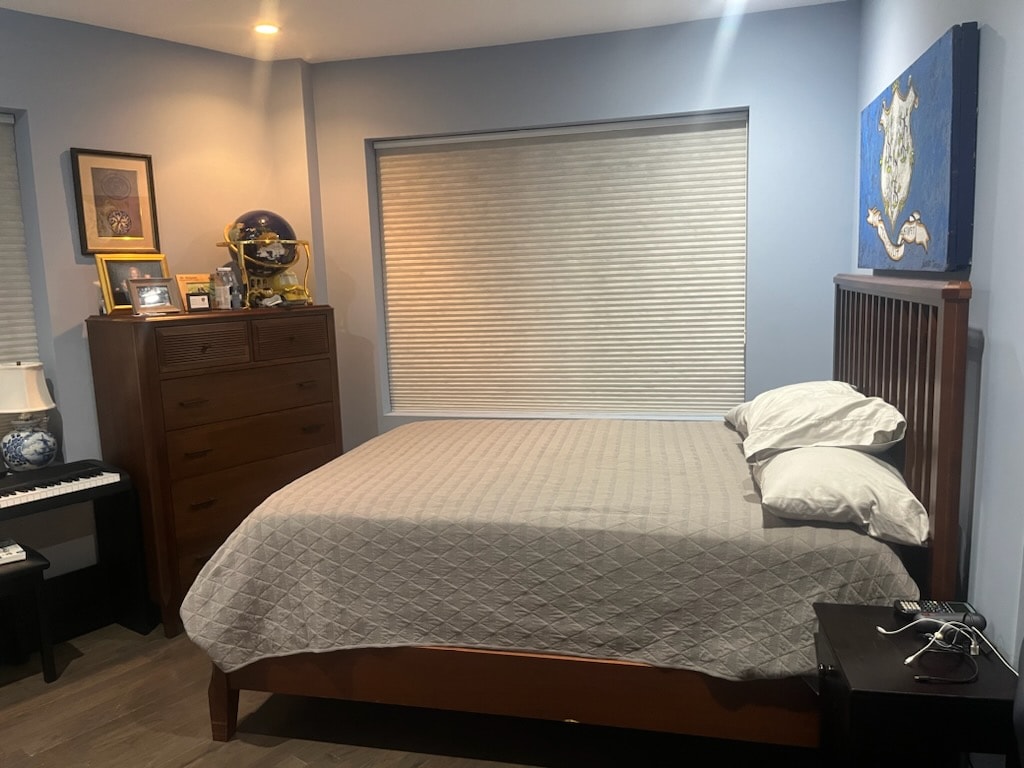 A cozy bedroom is highlighted by a spacious bed with neutral bedding and pillows. A wooden dresser stands against the wall, accompanied by a nightstand. A window with simple shades allows soft light to enter, complementing the calming blue walls.