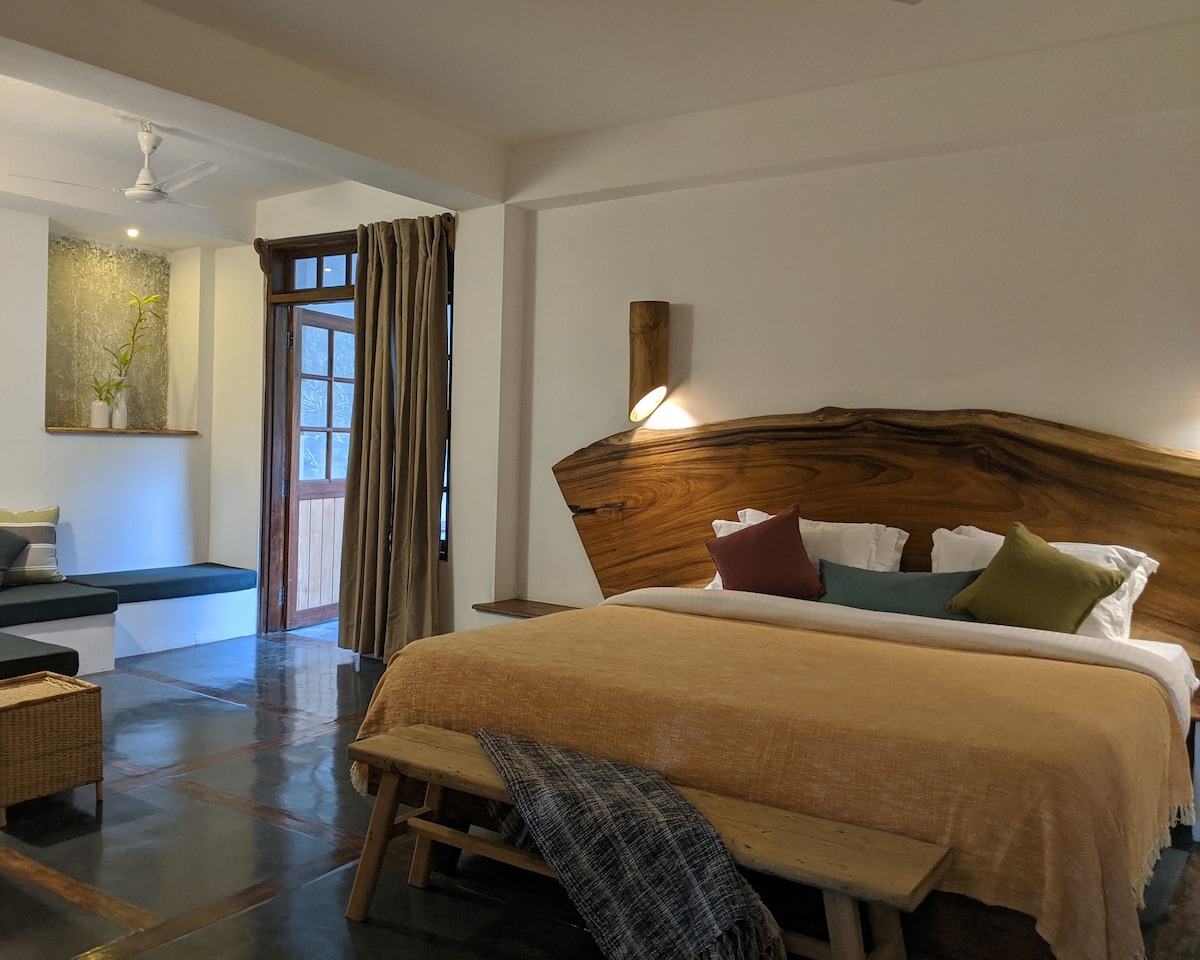 The spacious room features a large bed with a natural wood headboard, complemented by decorative pillows in various hues. A cozy seating area with a view is seen by the window, alongside a modest shelf displaying greenery. Warm lighting from wall fixtures enhances the ambiance.