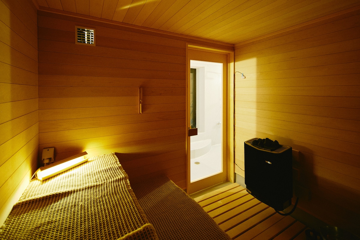 A serene sauna space is enclosed by wooden paneling, offering a calming glow from the light above. A flat bench, styled with textured fabric, runs along one side. A door leads to an adjacent area, while a small appliance sits on the opposite side.