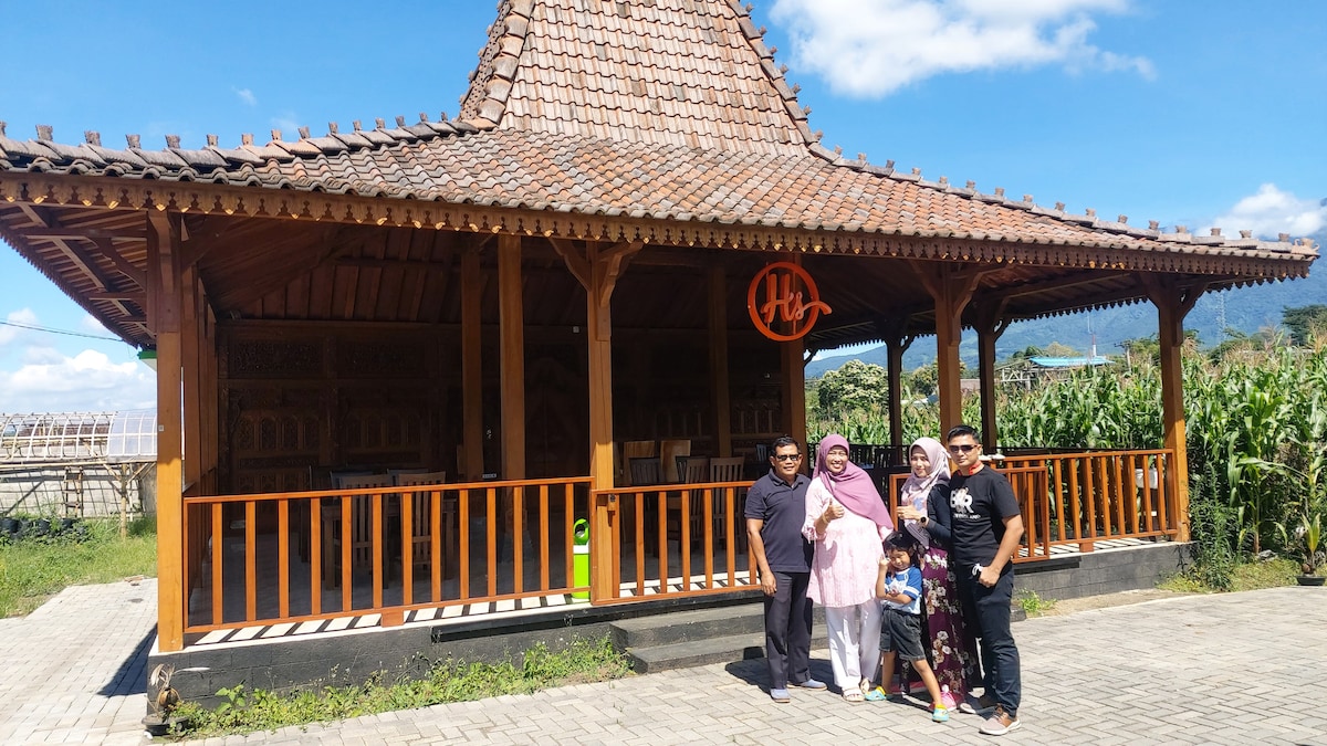 A traditional wooden pavilion features an intricately designed roof and a spacious porch. Surrounding greenery enhances the serene atmosphere. A family stands together in front, enjoying their time in this tranquil setting, with clear skies visible in the background.