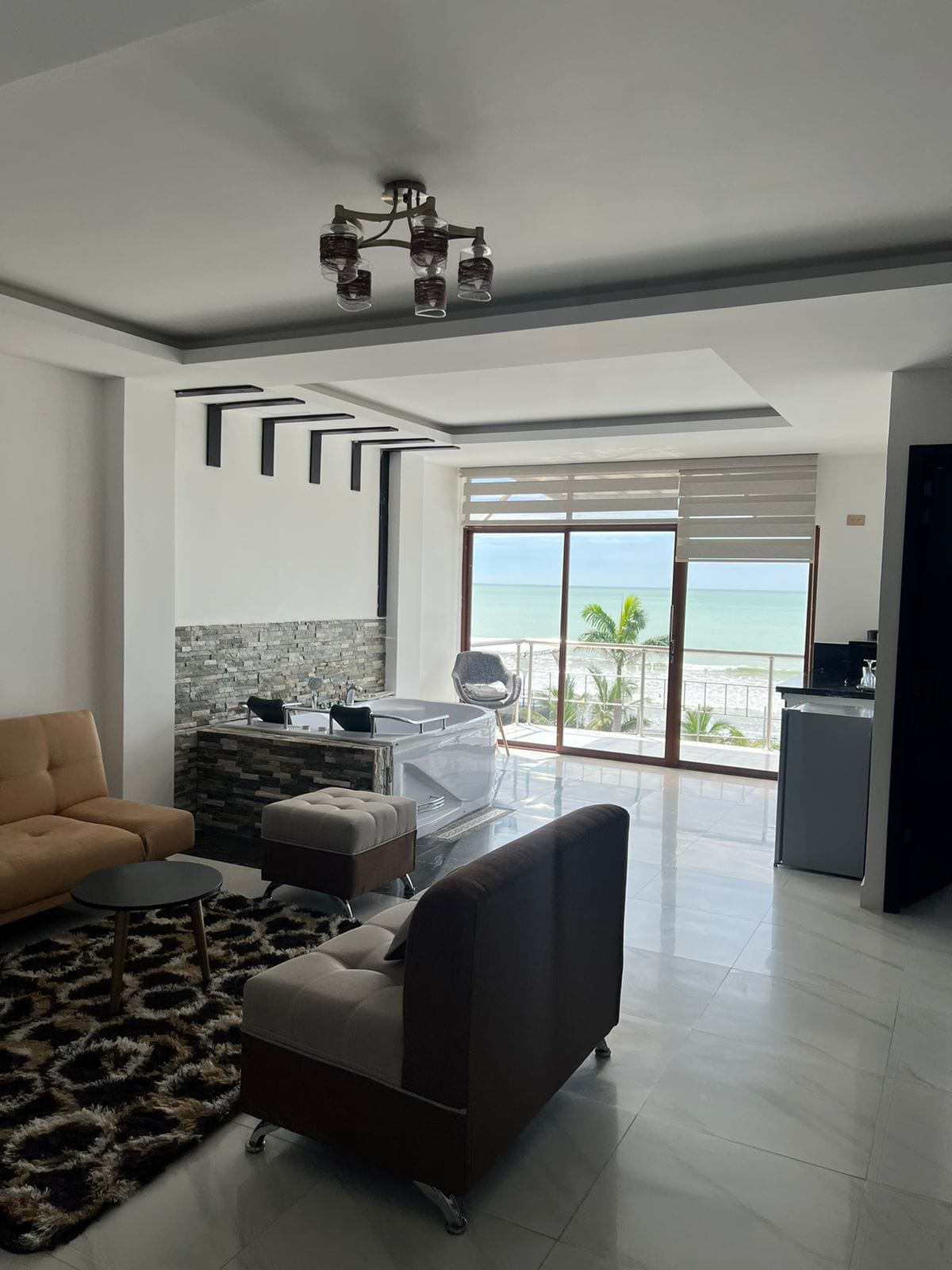 A modern living area features a light-colored sofa and two armchairs, arranged around a small table. Large windows provide views of the ocean, with sunlight illuminating the space. A textured stone accent wall adds visual interest, and a kitchenette is visible in the background.