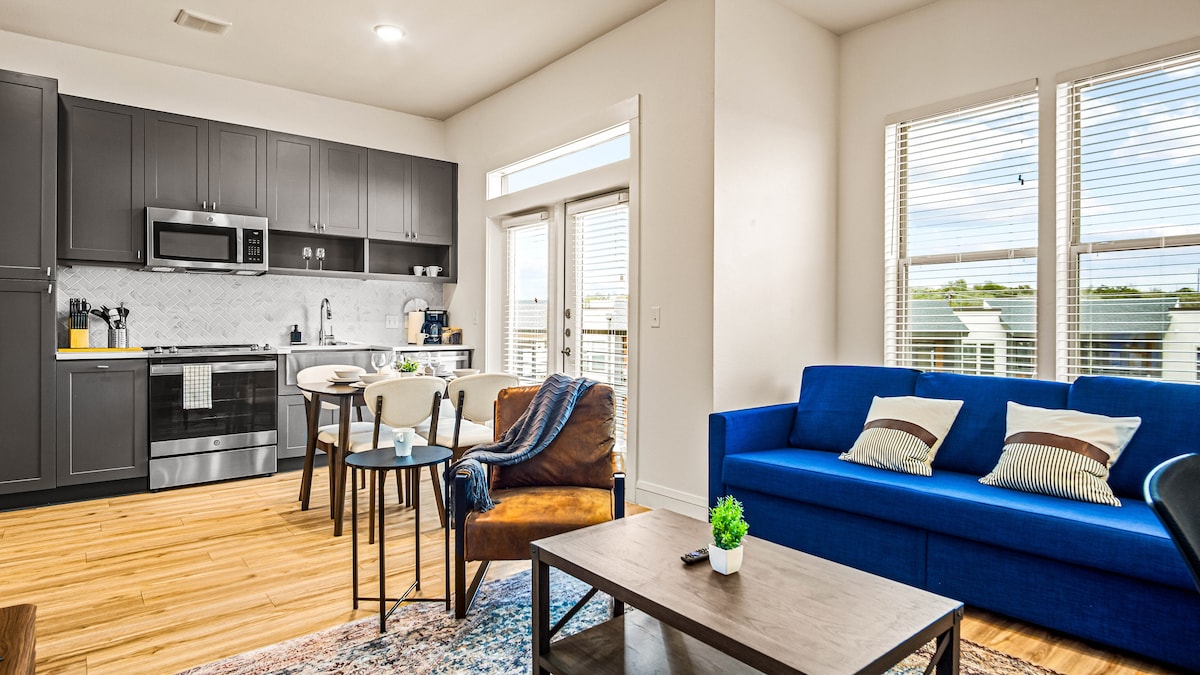 A modern open-plan living area features sleek cabinetry, a stainless-steel kitchen appliance set, and a dining table surrounded by chairs. A comfortable blue sofa with decorative pillows is positioned across from a coffee table, complemented by natural light streaming through large windows.