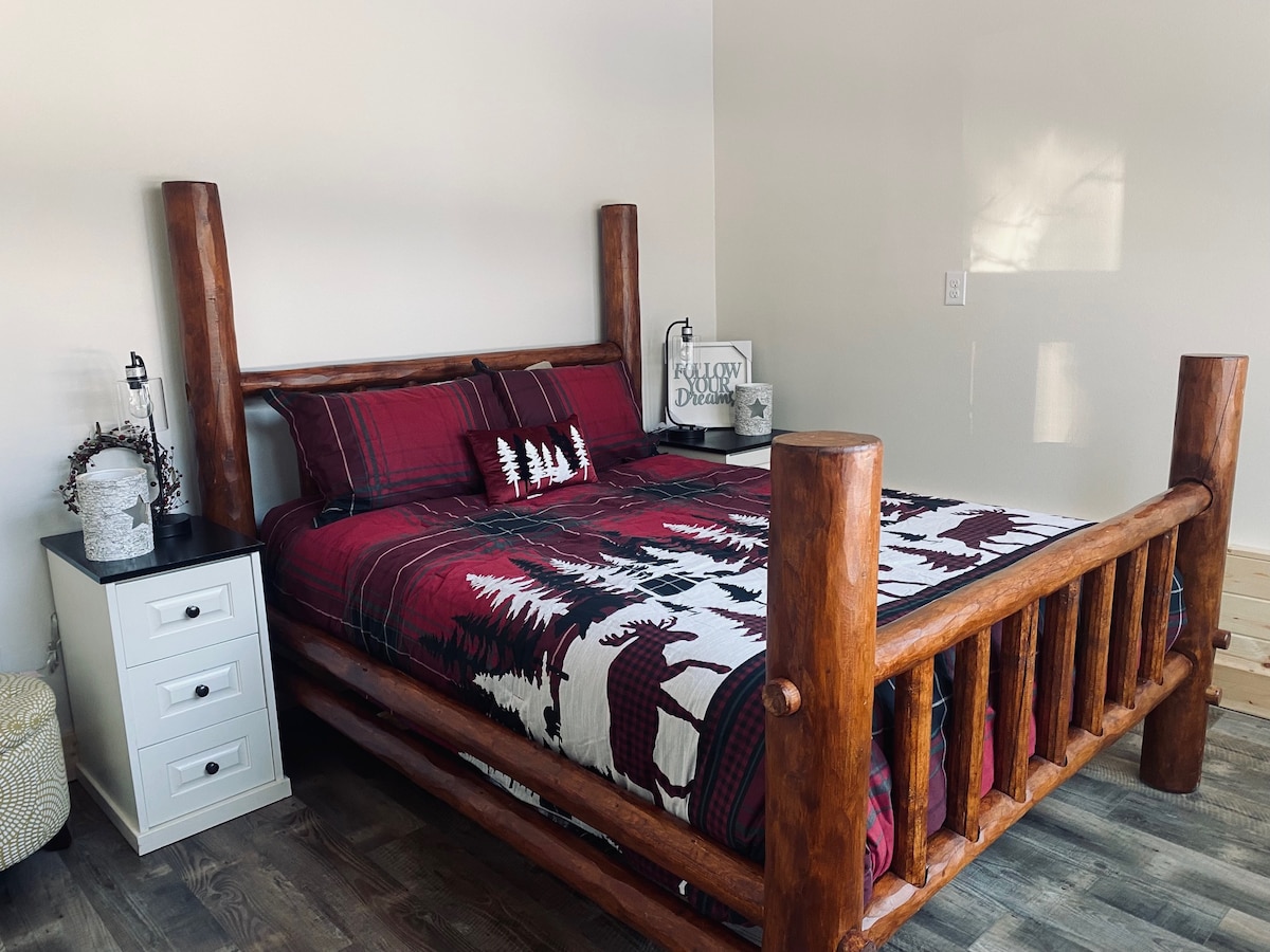 A rustic king bed crafted from wood logs features a patterned comforter with forest motifs. The bed is accompanied by a nightstand with a lamp and decorative accent. Warm light filters through the room, creating a cozy atmosphere.