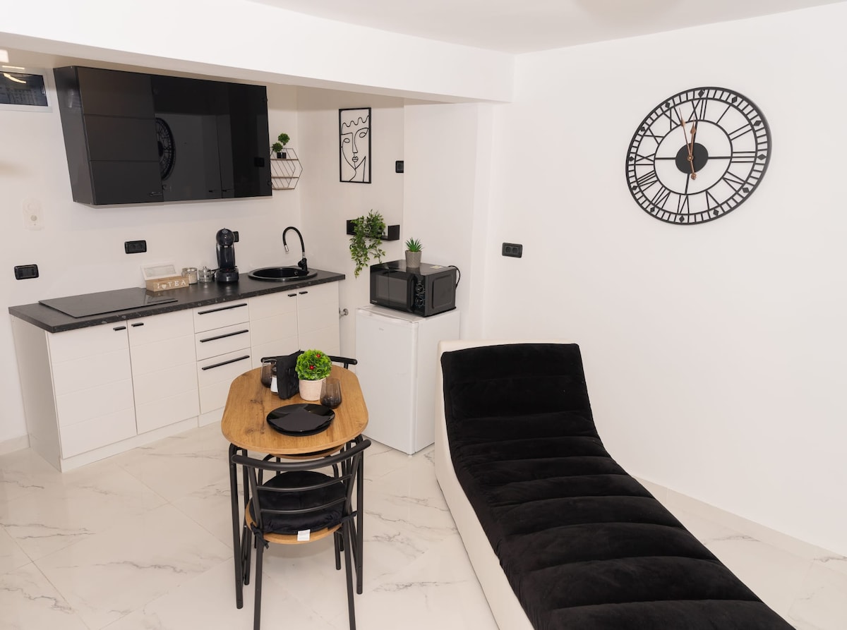 A modern kitchen area is visible, featuring sleek black cabinetry and a small dining table set for two. A black lounge chair is positioned nearby, enhancing the open layout. A wall clock and minimalistic artwork contribute to the contemporary decor.
