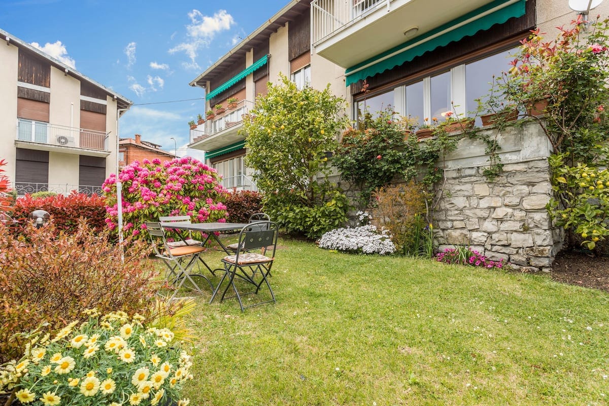 A well-maintained garden area features a grassy lawn surrounded by colorful flowering shrubs and plants. A small table with two chairs is positioned on the lawn, inviting outdoor dining. Nearby, vibrant yellow flowers add a cheerful touch to the serene environment.