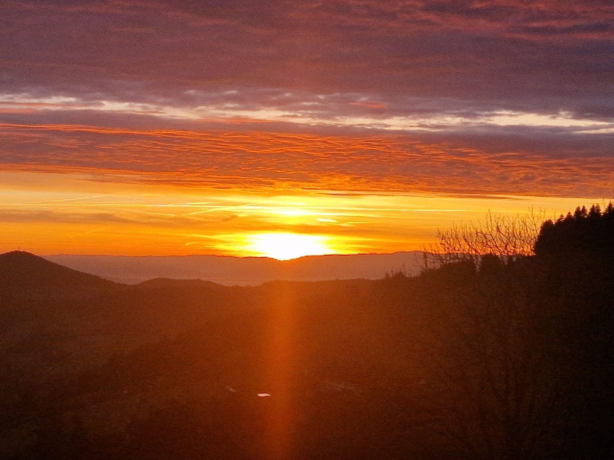 A vibrant sunrise is captured over distant mountains, casting warm hues of orange and gold across the sky. The rolling landscape is silhouetted against the light, creating a serene natural backdrop with streaks of clouds softly illuminated by the early morning sun.