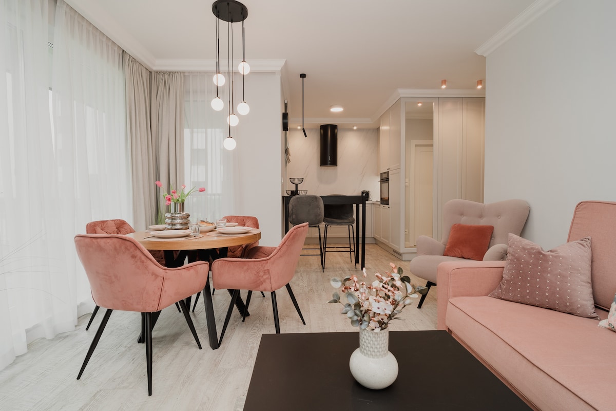 A modern dining area is highlighted by a round wooden table surrounded by upholstered chairs. Soft pink accents are present in the furniture, complemented by natural light streaming through sheer curtains. A small vase with flowers adds a touch of warmth to the space.