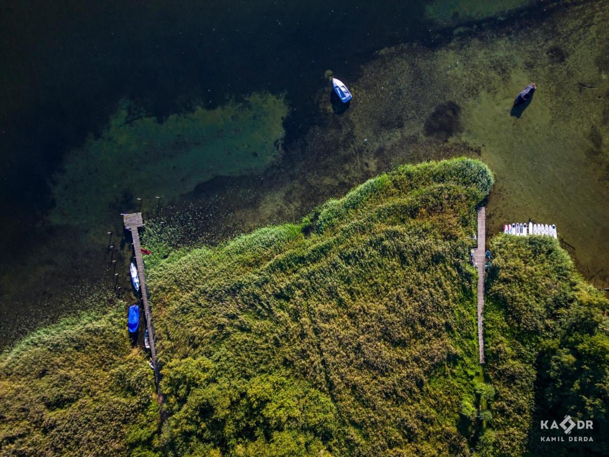 An aerial view captures the calm waters of Plauer See, with small boats gently floating near the shore. Lush greenery surrounds the water's edge, blending with wooden piers extending into the lake. The scene reflects a serene natural environment.