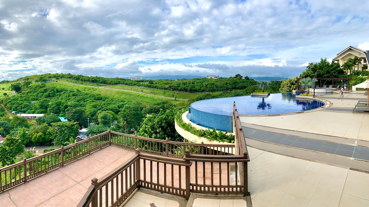 An expansive view showcases the infinity pool curving along the edge, reflecting the serene sky above. Lush green hills stretch into the distance, while a spacious terrace with wooden railings offers a place for relaxation. The landscape blends nature with the tranquil water.
