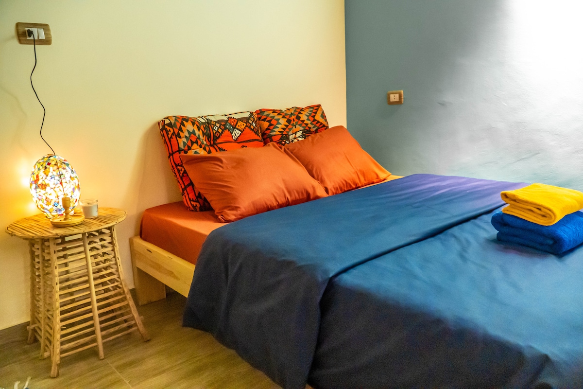 A spacious bed is dressed in contrasting linens featuring vibrant orange and deep blue tones. A woven side table holds a decorative lamp, casting a warm light. Additional towels in bright colors are neatly placed at the foot of the bed, enhancing the inviting feel of the room.