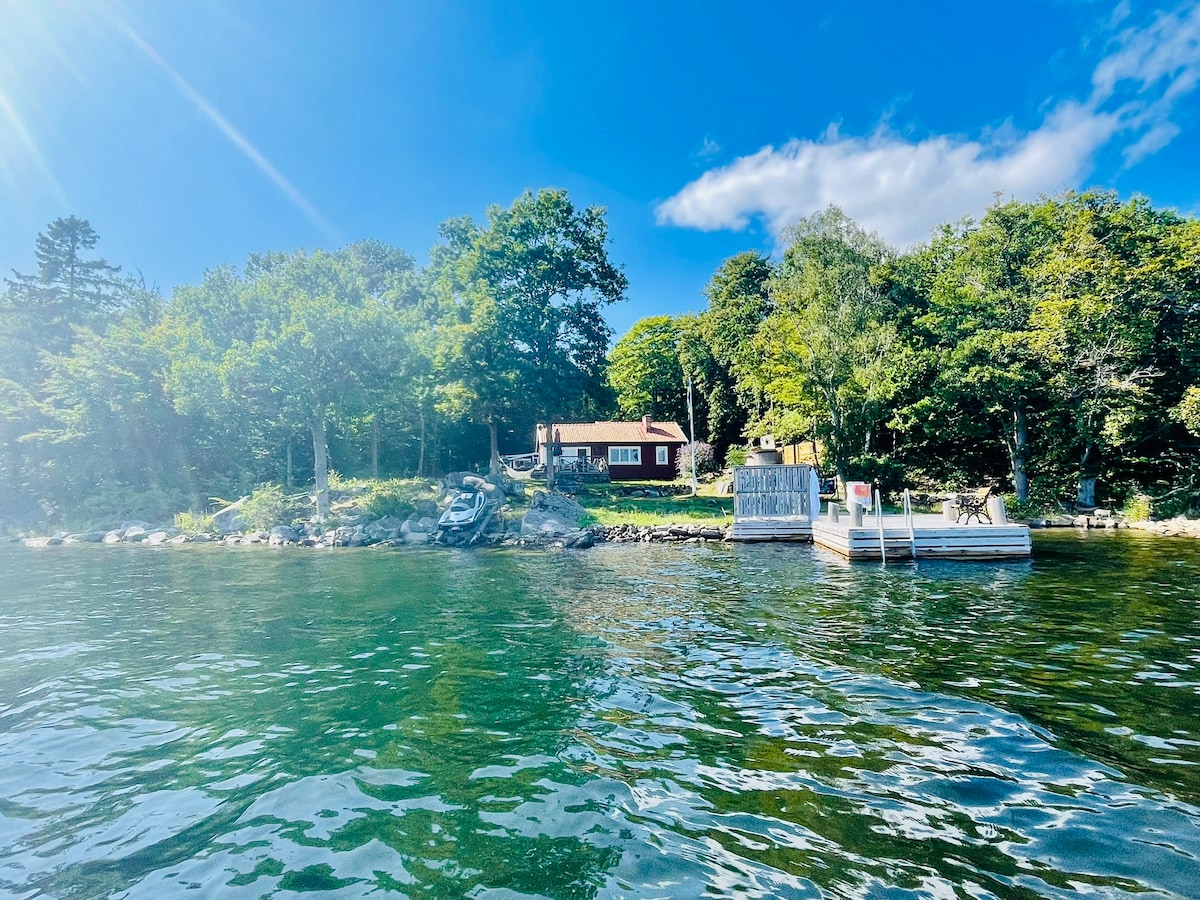 The view showcases a charming red holiday house nestled among lush trees, positioned on a waterfront. A wooden dock is visible, providing access to the water. The surrounding landscape features rocky outcrops and vibrant greenery under a clear blue sky.