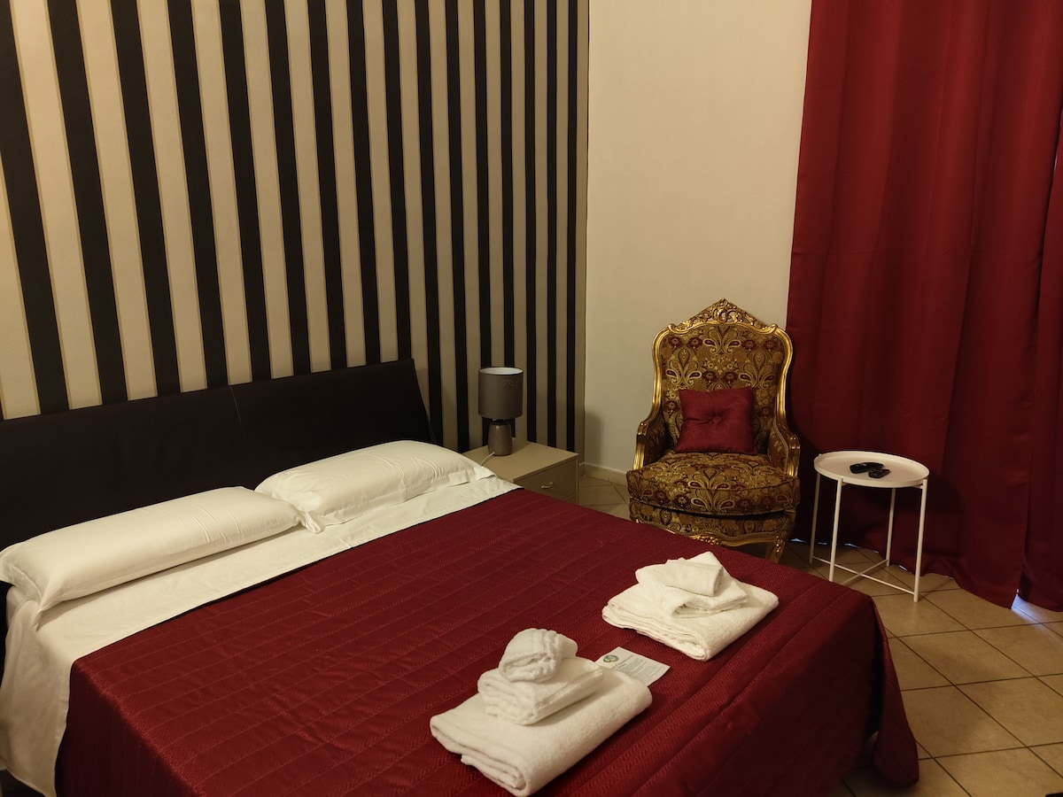 A cozy room features a spacious bed covered with a red quilt, complemented by neatly arranged towels. A decorative armchair in a bold pattern sits beside a small white table. A vibrant red curtain frames the window, while striped wallpaper adds texture to the walls.