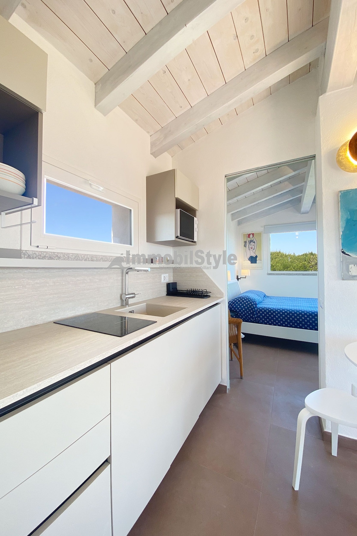 A compact kitchen area features modern cabinetry, a sink, and ample countertop space. Directly ahead, a cozy sleeping area can be seen through an arched opening, framed by a bright blue wall. Natural light fills the space from a nearby window.
