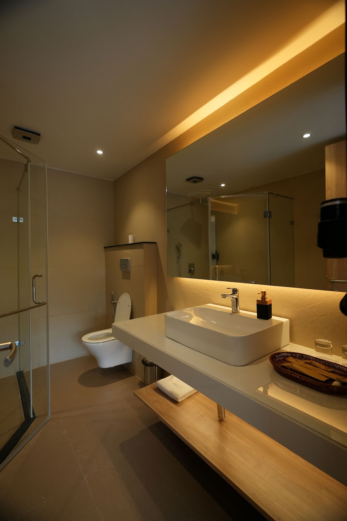 A modern bathroom features a spacious shower area enclosed in glass. A sleek white sink sits atop a minimalist countertop, illuminated by warm lighting. The mirror reflects a clean design, enhanced by neutral tones and wooden accents.