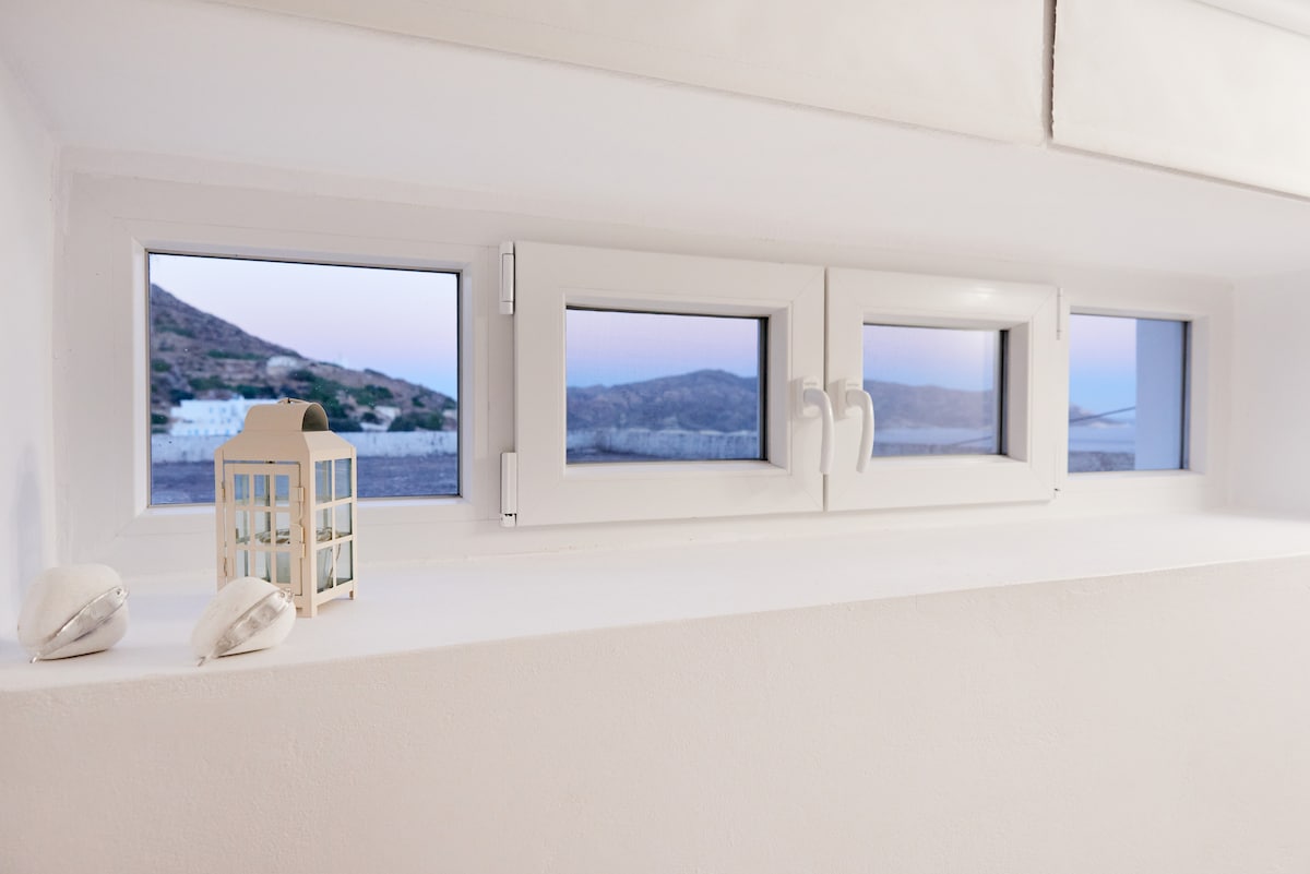 A row of three windows is visible, framed in white and offering a panoramic view of the surrounding landscape. A small lantern is placed beside two seashells on the windowsill, enhancing the coastal ambiance.