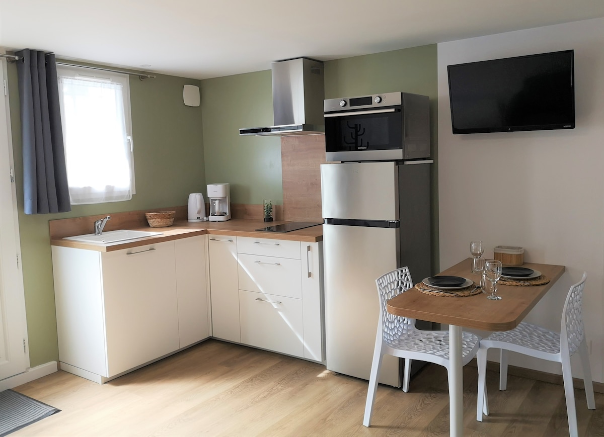 The kitchenette features modern appliances, including a compact refrigerator and microwave. A small dining area with a wooden table and two white chairs is positioned under a wall-mounted television. Natural light enters through a window, complemented by gray curtains.
