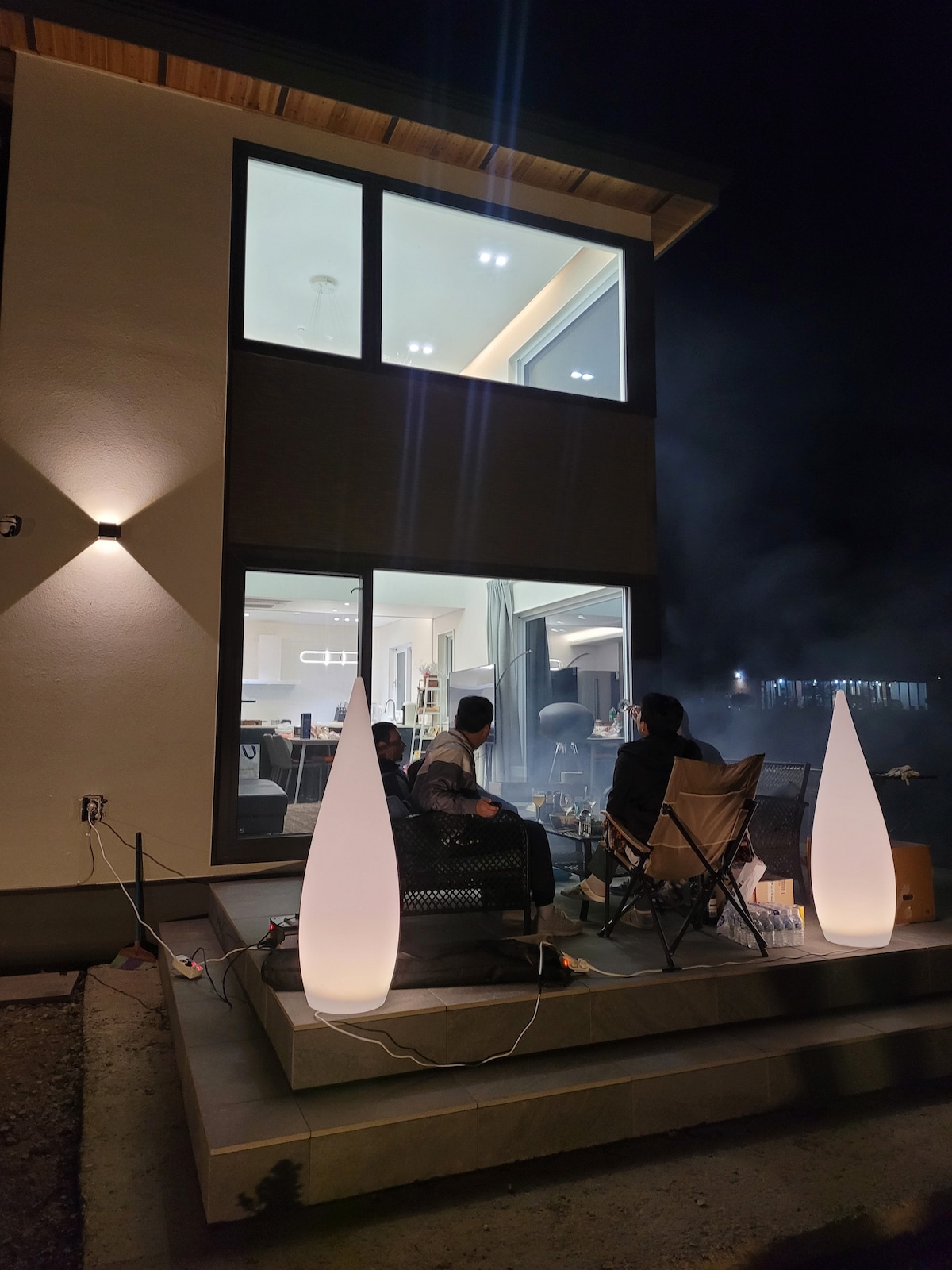 An inviting outdoor space is shown at night, featuring illuminated, sculptural light fixtures flanking a seating area. The soft glow enhances the ambience as guests gather around a fire pit, with the modern structure and large windows of the house visible in the background.