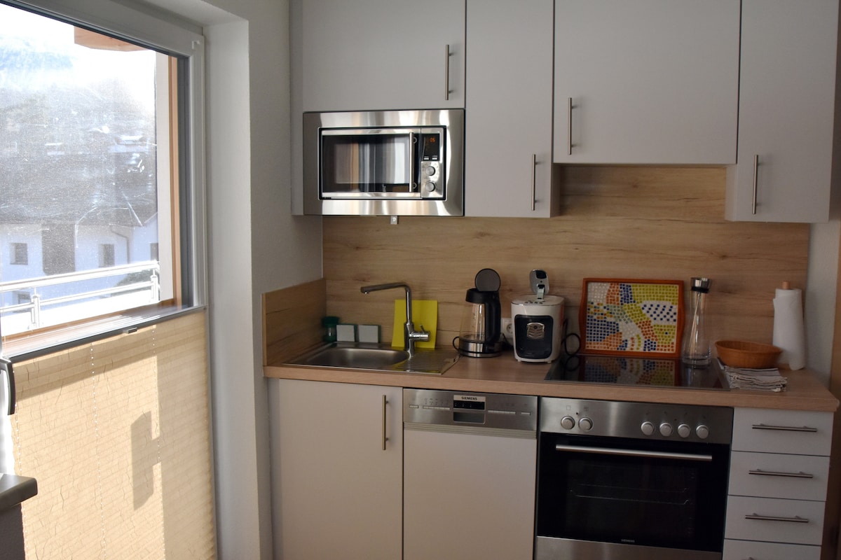 The compact kitchen features modern appliances, including a microwave, oven, and coffee maker. A sink is positioned beneath a window, allowing natural light to illuminate the wooden countertop. Neutral cabinetry complements the functional layout, with hints of color from a wall art piece.