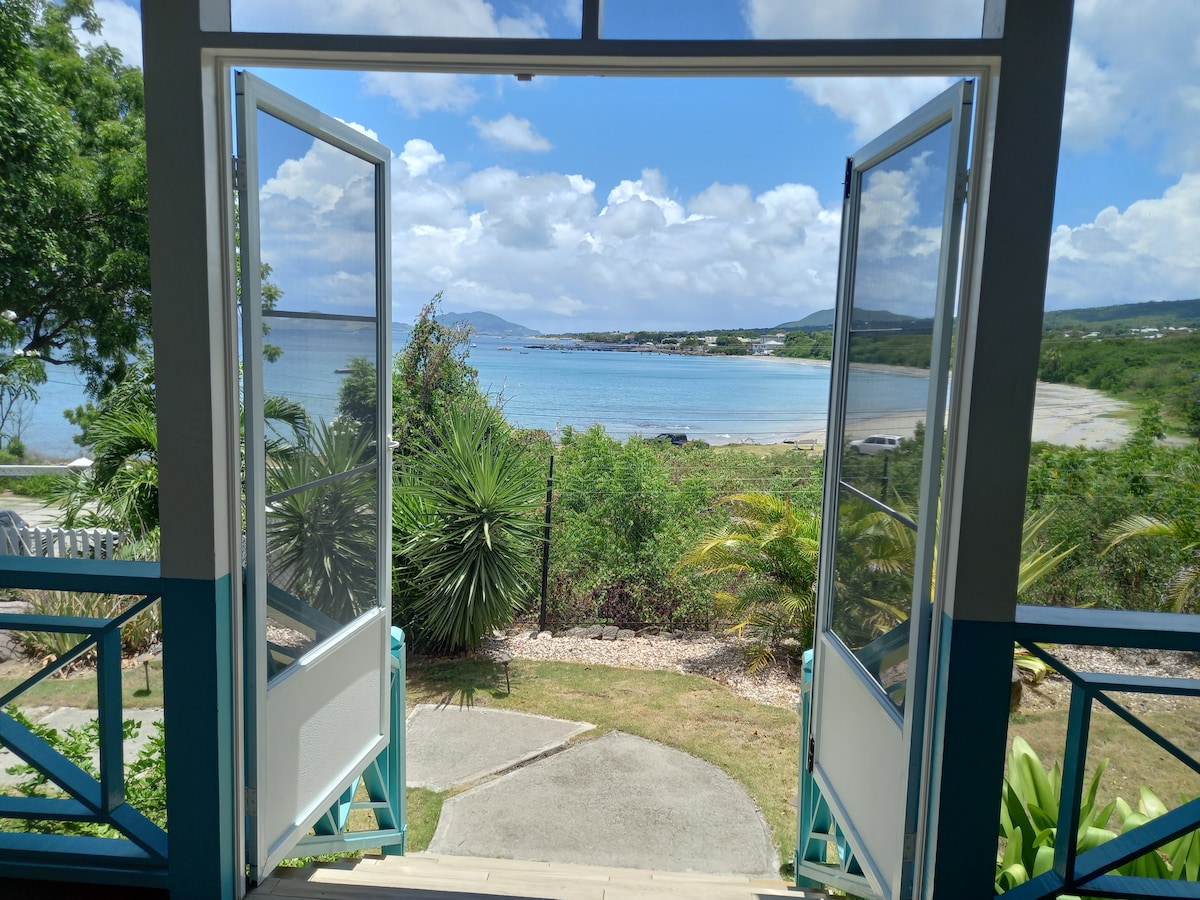 Double French doors open onto a scenic view of Gallows Bay, framed by lush greenery. A pathway leads to the beach, with gentle waves visible in the distance under a partly cloudy sky.