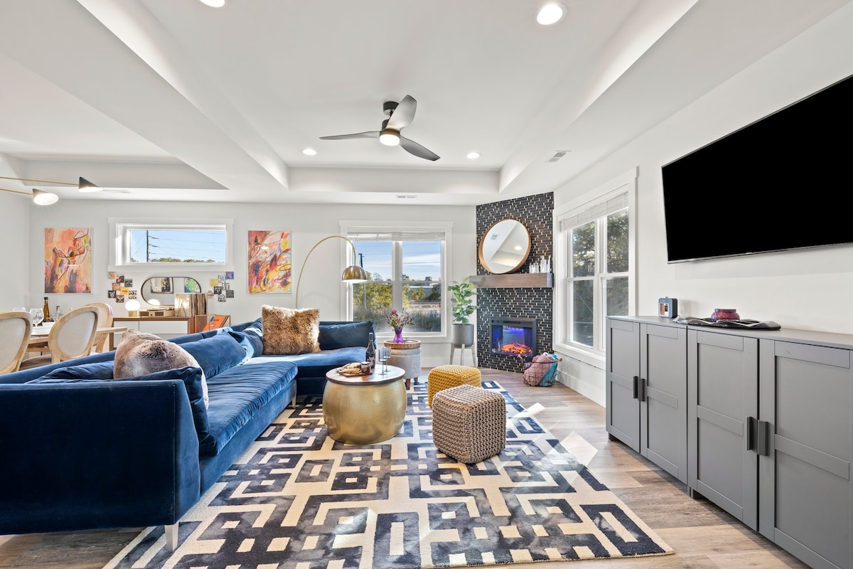 A spacious living area features a blue sectional sofa and a large round coffee table on a geometric patterned rug. Natural light fills the room through multiple windows, and a smart TV is mounted on the wall opposite a cozy electric fireplace.