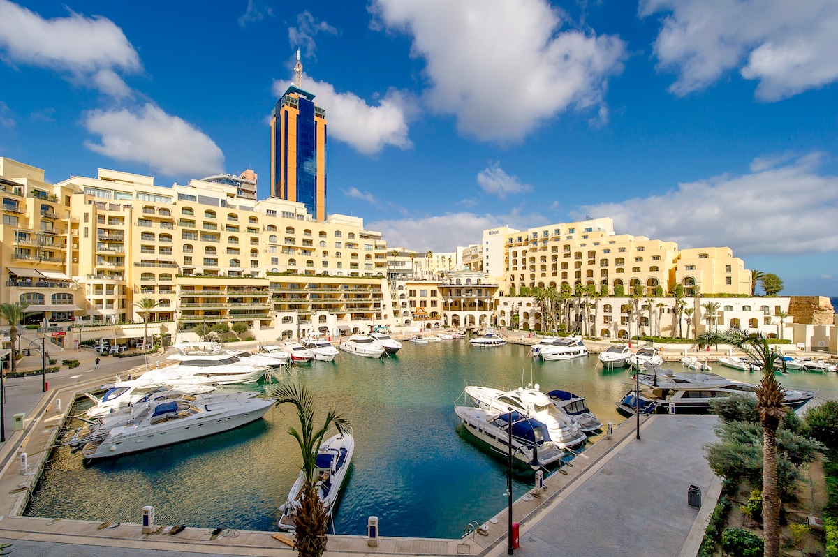 A vibrant marina is featured, surrounded by modern buildings and palm trees. Several yachts are docked along the water's edge, reflecting the clear blue sky. The scene captures a lively yet serene atmosphere with the city's architectural beauty in the background.