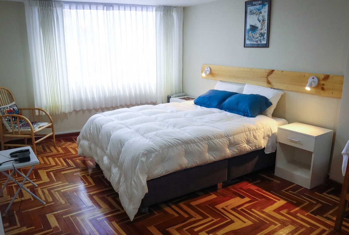 A clean bedroom features a spacious bed with a white comforter and blue accent pillows. A rattan chair sits beside a small side table. Natural light enters through large, sheer-curtained windows, illuminating the parquet floor.