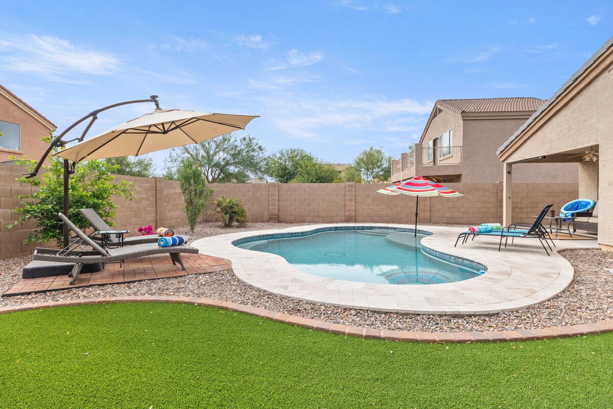 A private pool is surrounded by a manicured lawn and decorative gravel. Comfortable lounge chairs are positioned nearby, with colorful pool floaties and an umbrella providing shade. A neutral-colored home is visible in the background, framed by desert landscaping.