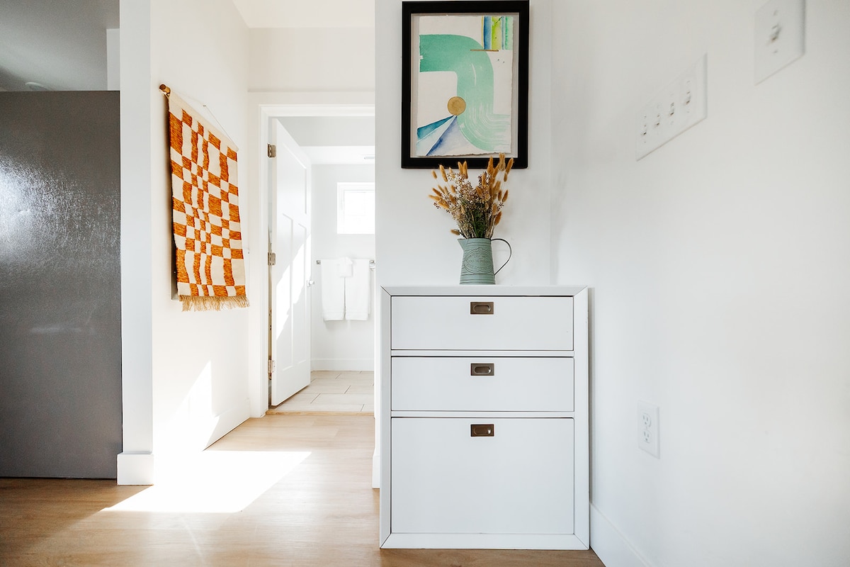 A bright entryway features a small dresser with three drawers, topped with a vase holding dried flowers. A piece of abstract art hangs above the dresser. A colorful woven wall hanging adds texture, with a door leading to a bathroom visible in the background.
