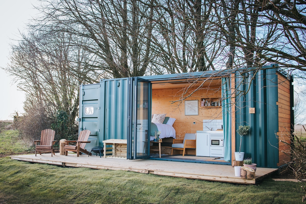 A modern tiny cabin constructed from a shipping container features open sliding doors, allowing natural light to fill the space. A spacious bed is positioned inside, complemented by a kitchenette and minimalistic decor. The outdoor deck includes two wooden chairs, offering a serene setting surrounded by greenery.