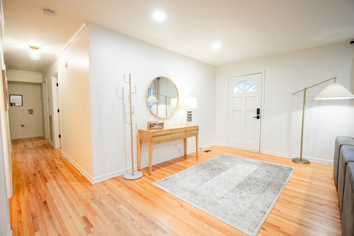 An inviting entryway features warm hardwood floors and a soft area rug. A round mirror hangs above a light wooden console table, complemented by a floor lamp. A door with a window provides natural light, leading to a spacious corridor.