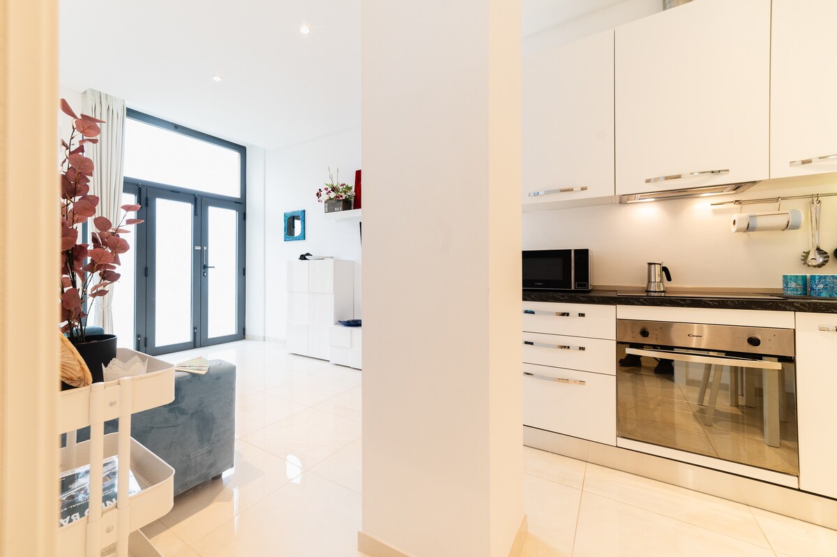 A modern living space is shown featuring a kitchen with sleek cabinets and stainless steel appliances. Natural light floods the area through large glass doors, while decorative plants add a touch of warmth to the minimalistic decor. The tiled floor reflects the light, enhancing the spacious feel.