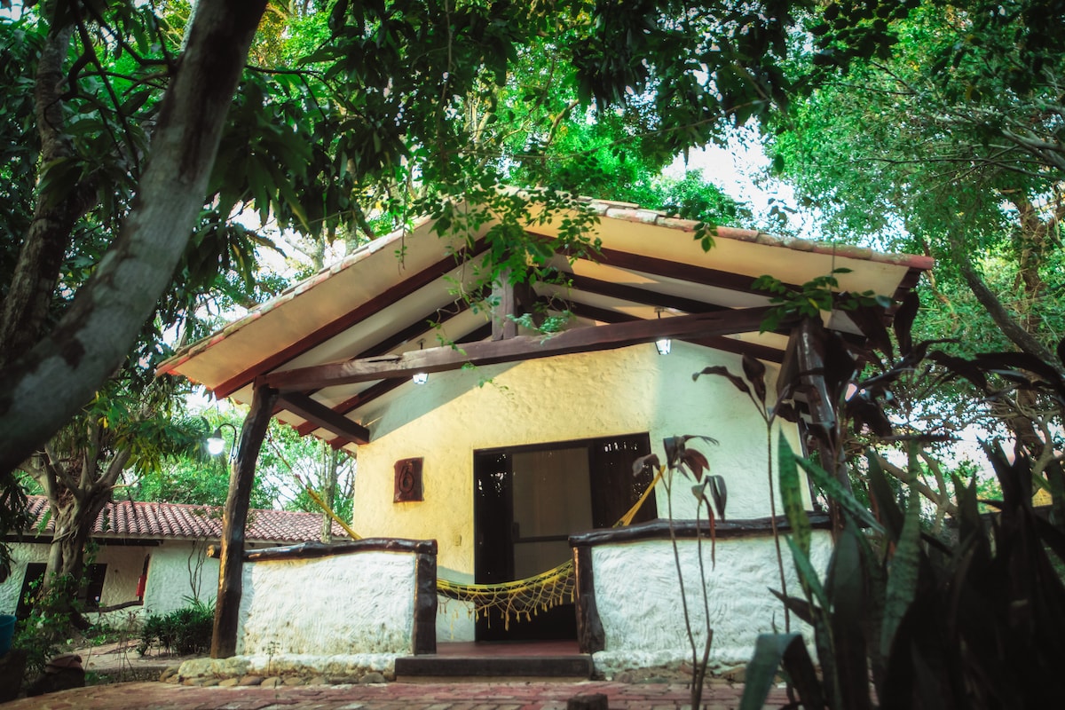 A rustic cabin is nestled among lush greenery, complete with a sloped roof and wooden beams. The entrance features a spacious porch that is adorned with plants and lights, inviting relaxation in a natural setting.