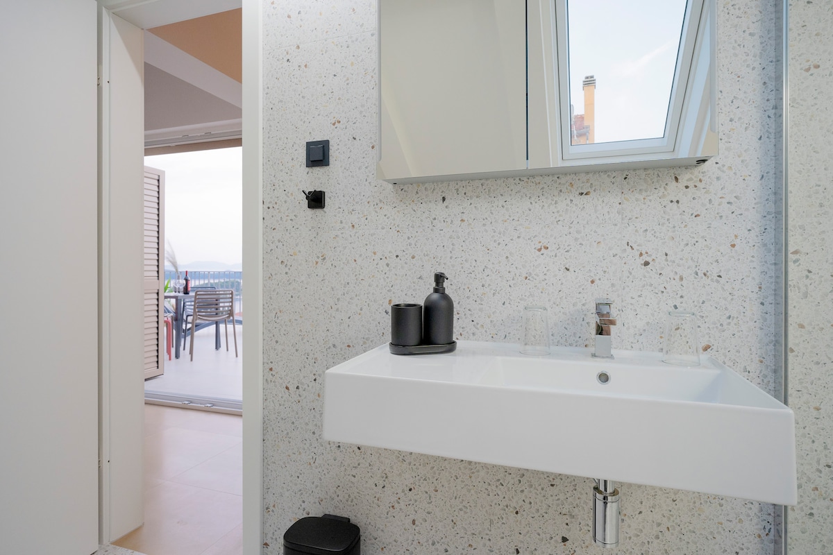 A modern bathroom features a sleek, wall-mounted sink with a minimalist design. Natural light enters through a skylight, illuminating the textured walls. A doorway leads to an outdoor area, suggesting a connection to the private balcony with additional seating.