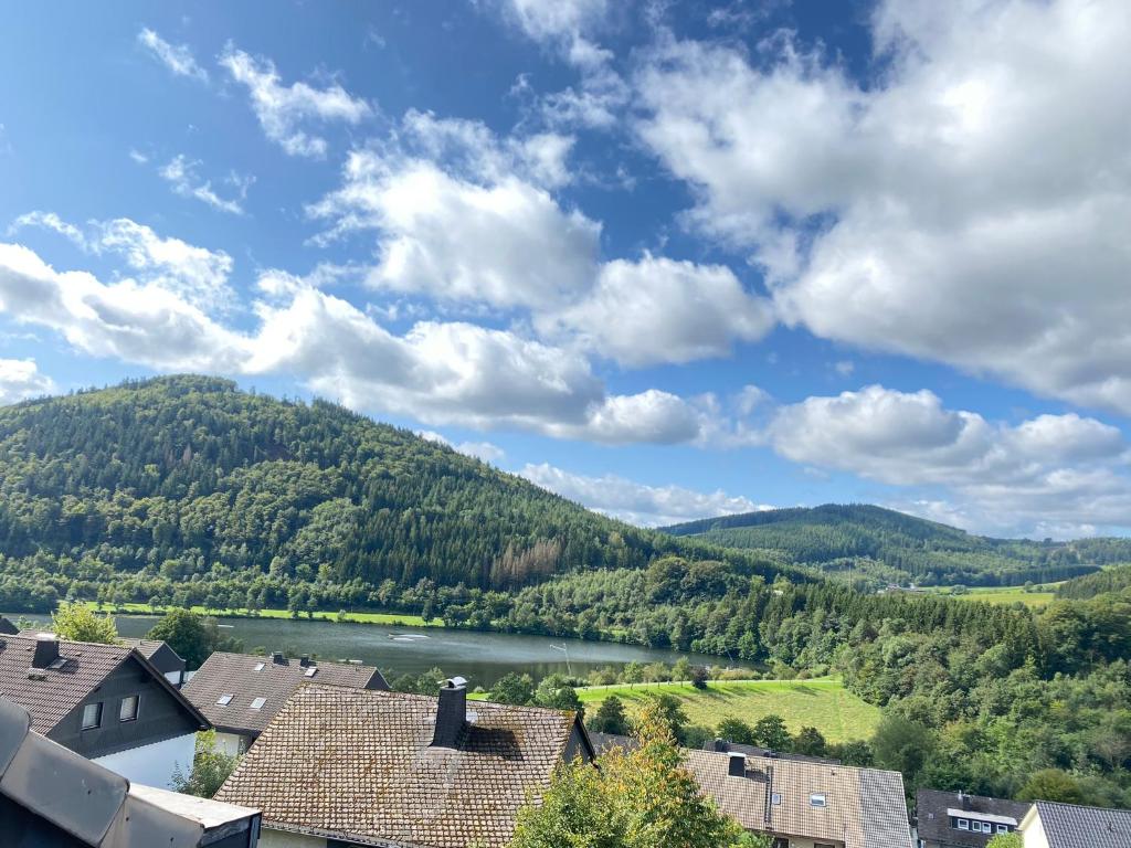 A panoramic view captures rolling green hills surrounding a serene lake, reflecting the blue sky dotted with fluffy clouds. A cluster of rooftops is visible in the foreground, leading the eye towards the tranquil waters and lush landscapes beyond.