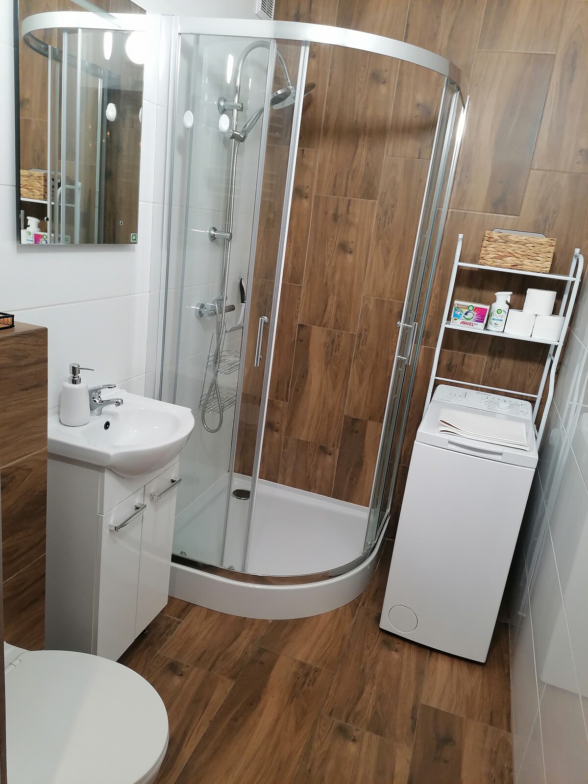 The bathroom features a glass-enclosed shower, a compact sink, and a toilet. A laundry unit is positioned next to a shelf that holds toiletries. Warm wood paneling enhances the modern design, while mirrors reflect light and create a sense of openness.