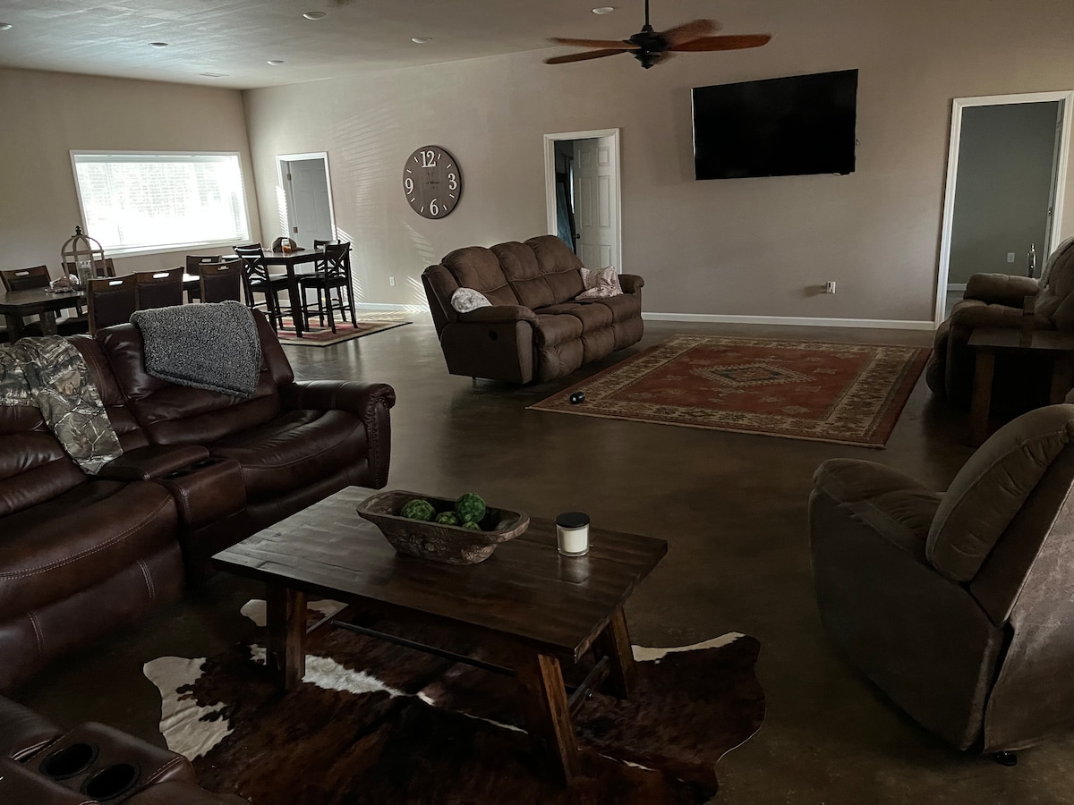 A spacious living area features a large rug atop a concrete floor, with comfortable brown seating arranged around a wooden coffee table. A wall-mounted television and large wall clock are visible, and natural light fills the room through multiple windows.