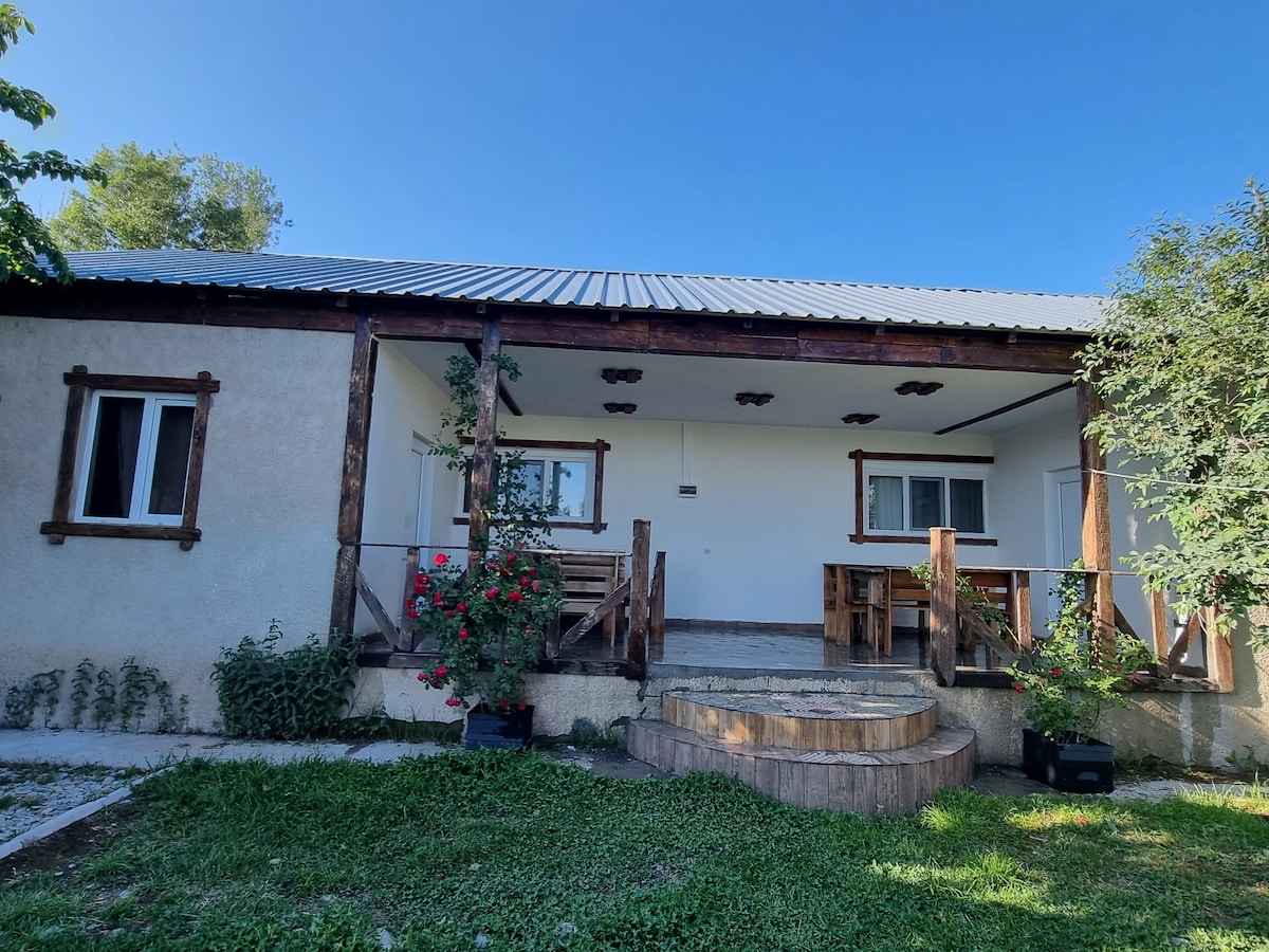 A guesthouse exterior is visible, featuring a wooden deck with two benches. Surrounding greenery includes shrubs and flowers, adding a touch of color. The building has a simple façade with two windows and a sloped roof, all set against a clear blue sky.